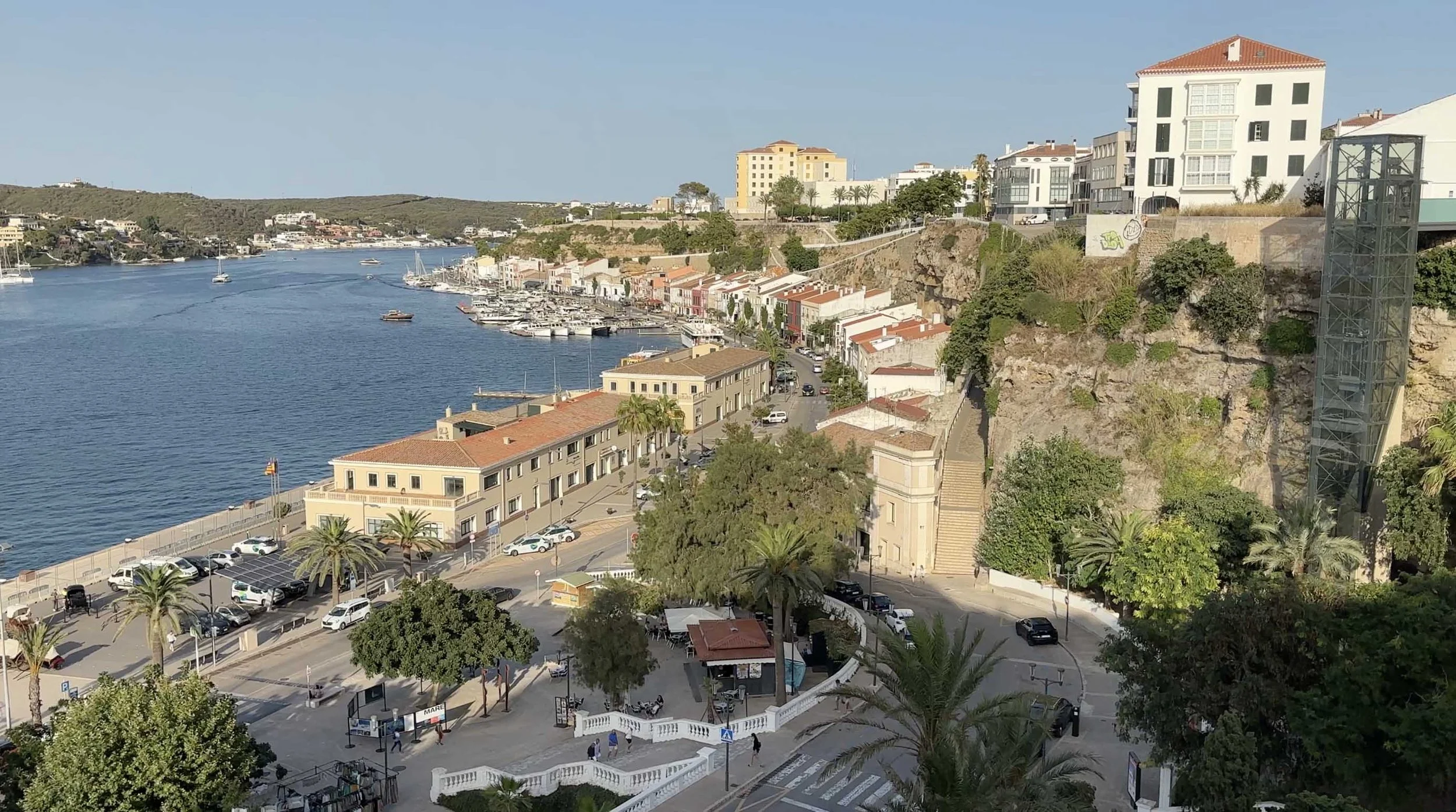 View over a harbor in Menorca, Spain, with moored boats, waterfront buildings and palm trees below a rocky hillside.