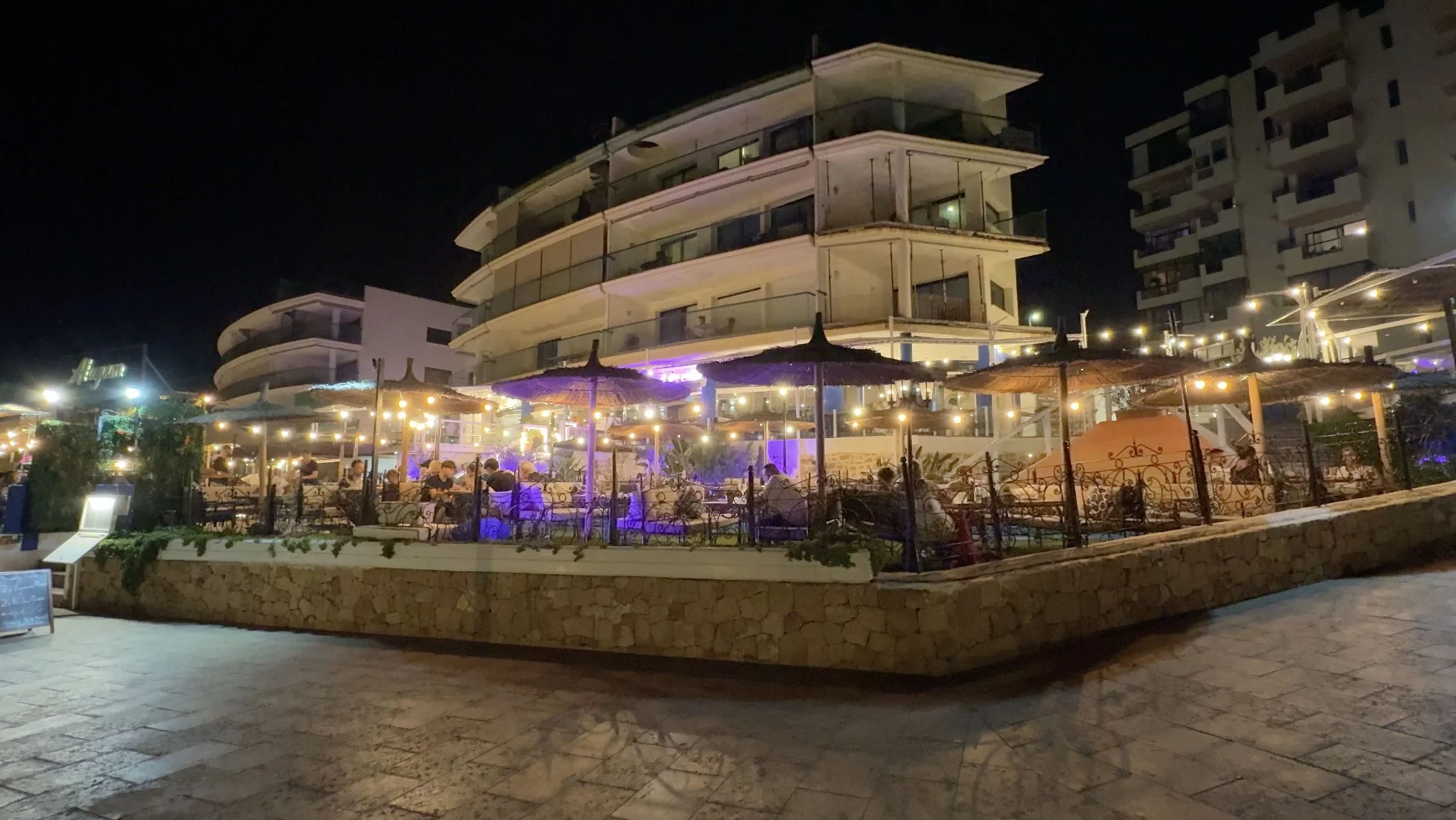 Diners on an outdoor restaurant terrace in San Antonio, Ibiza, beneath straw parasols and string lights at night.