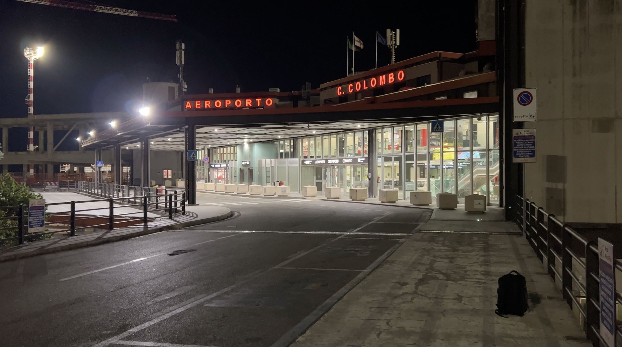 Genoa Airport entrance at night with illuminated red lettering and a backpack on the pavement.