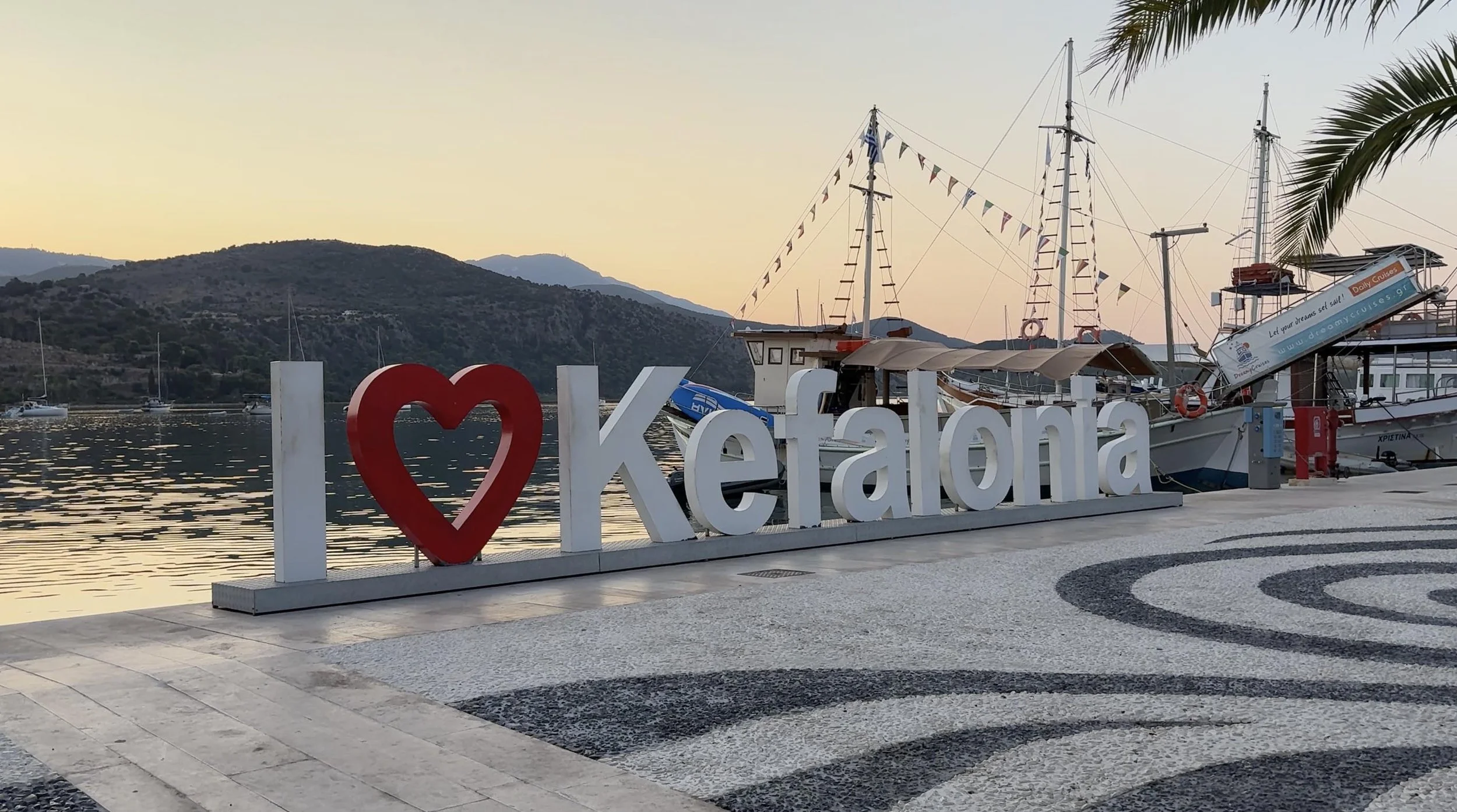 “I love Kefalonia” sign on the waterfront in Kefalonia, Greece, with boats and hills at sunset.