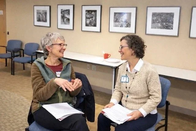 Two women sit facing each other with papers in their laps, smiling.