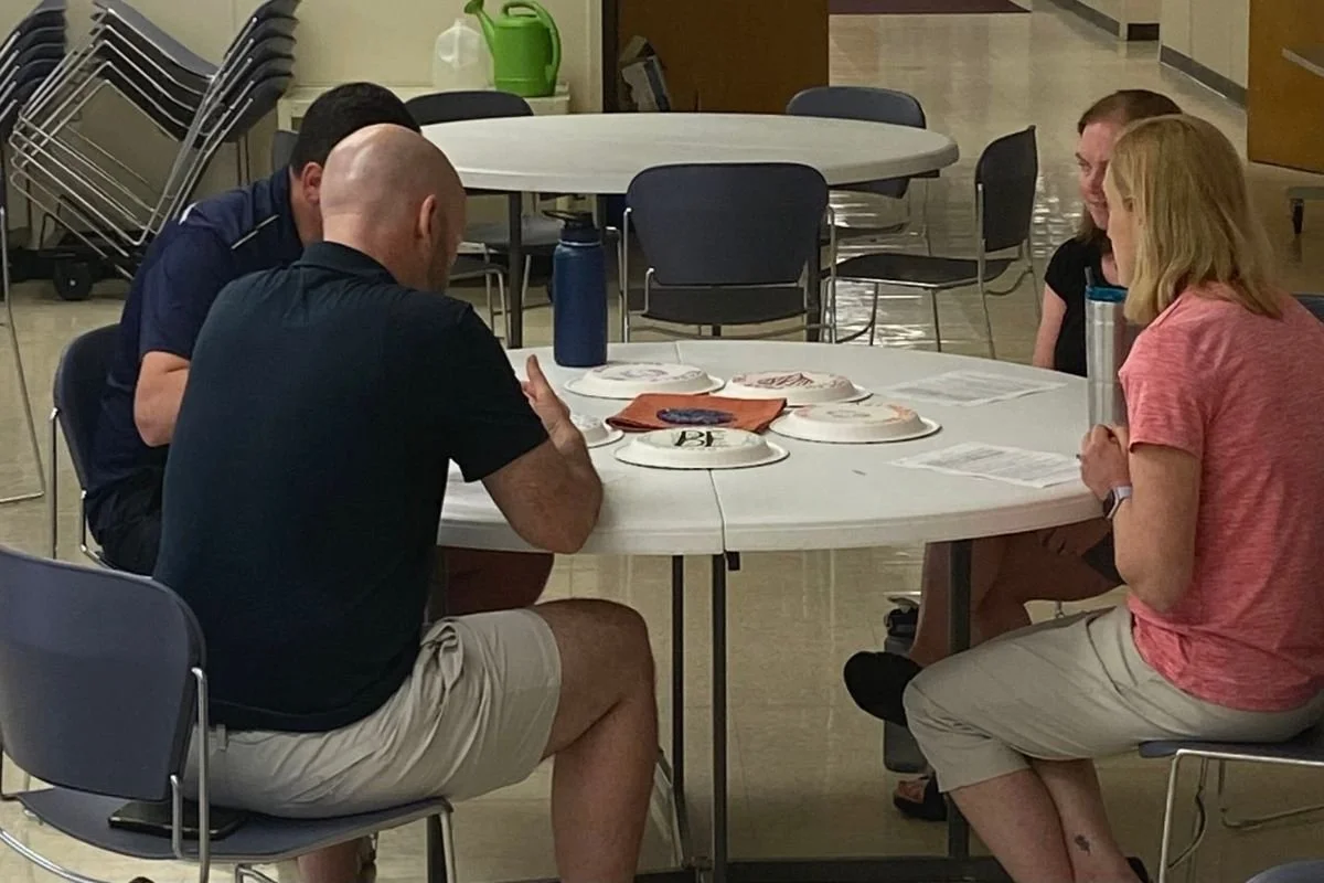 Two white men and two white women sit in blue chairs around a white circular table. There is an orange cloth surrounded by five paper plate with writing in the center of the table.