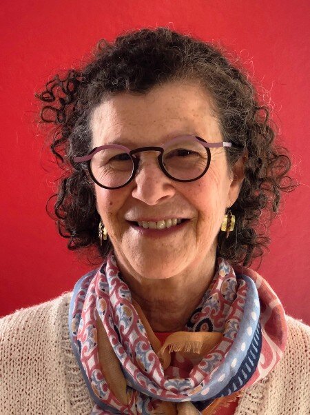 Older white woman with curly hair and round framed glasses in front of a red background.