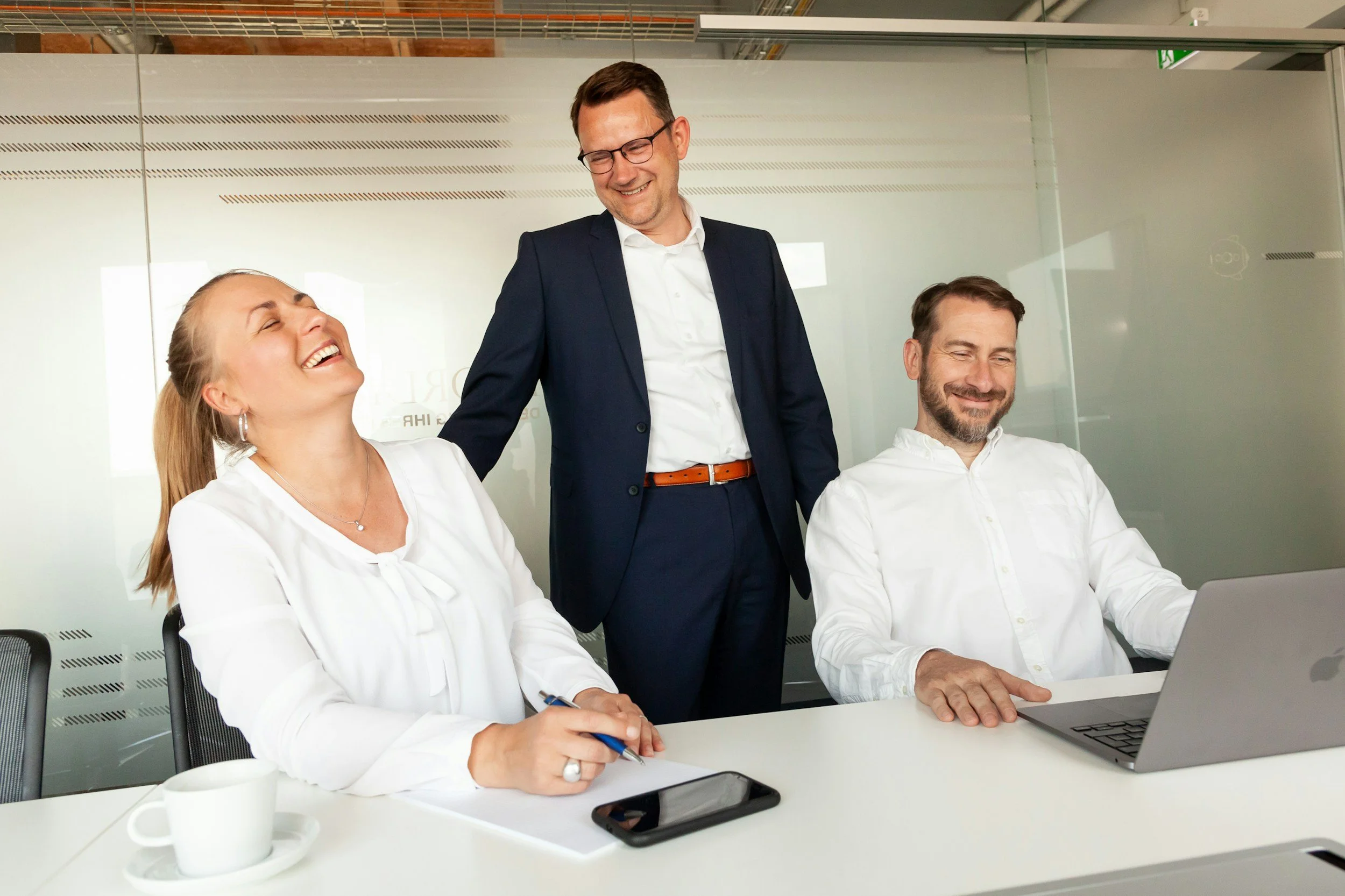 Two employees, in white, sit at a desk while their supervisor stands behind them in a dark suit. All are smiling.