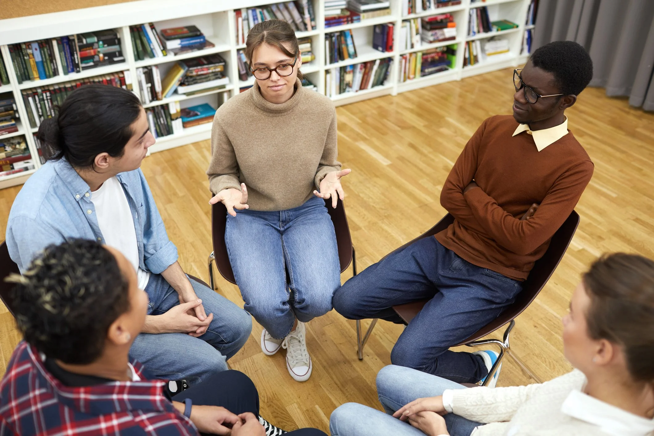 A diverse group of six young adults sitting in a circle, engaged in a discussion in a room with bookshelves.