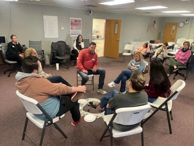 Teachers and administrators sit in a circle with a centerpiece in a carpeted meeting room. Other participants sit in another circle behind them, observing, in a "Fishbowl" role play.