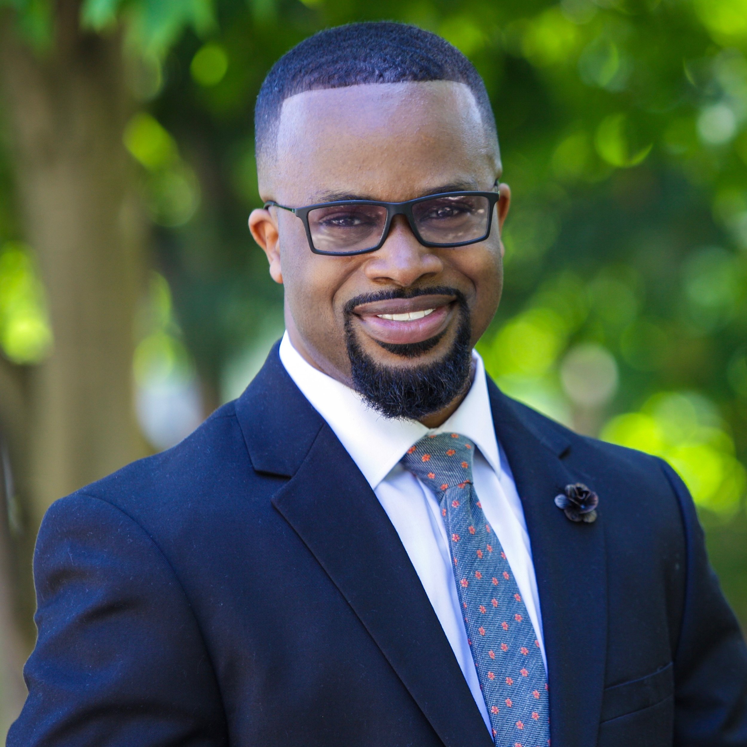 A black man with a goatee wearing black rimmed glasses, white shirt, blue suit jacket and light blue tie.