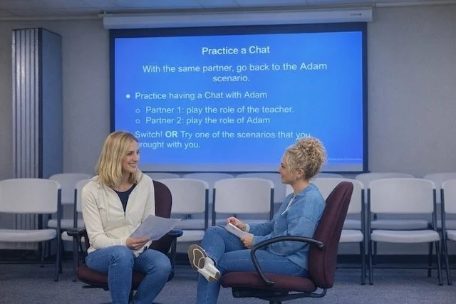 Two women sit facing each other as they practice a role play in front of a blue screen