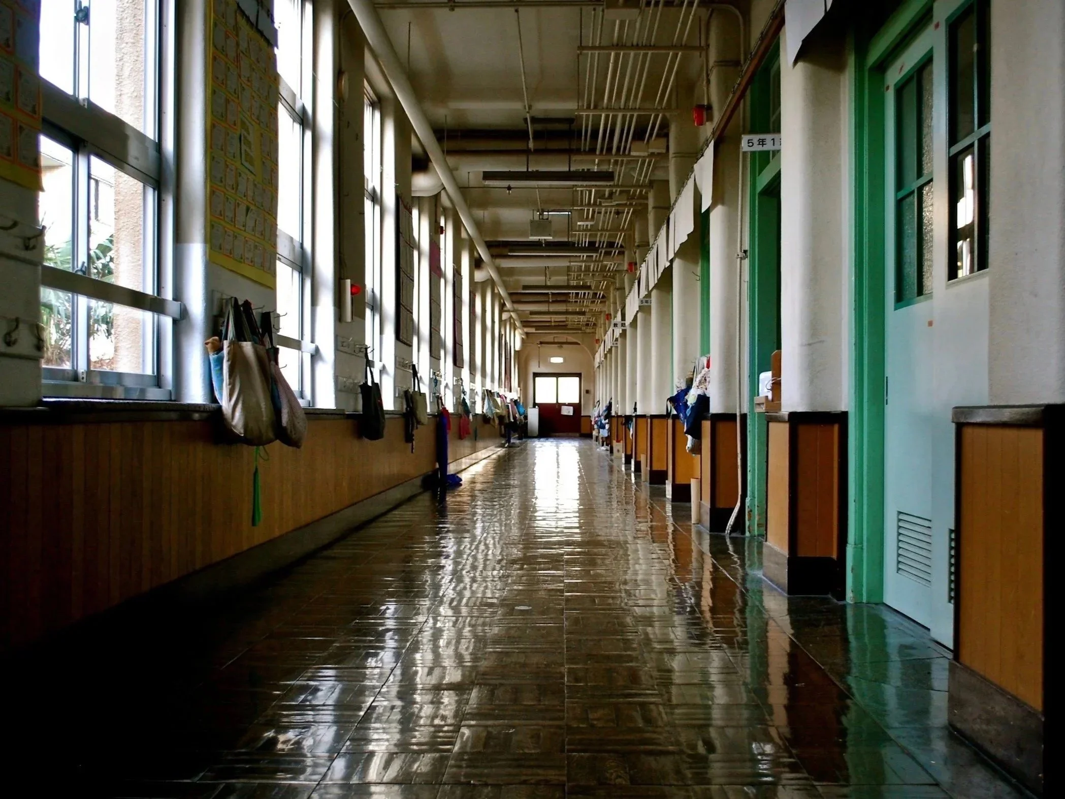 long school hallway with a shiny floor and double doors at the end. Windows on the left side of the hall with backpacks hanging between them. Classroom doors on the right side of the hall.