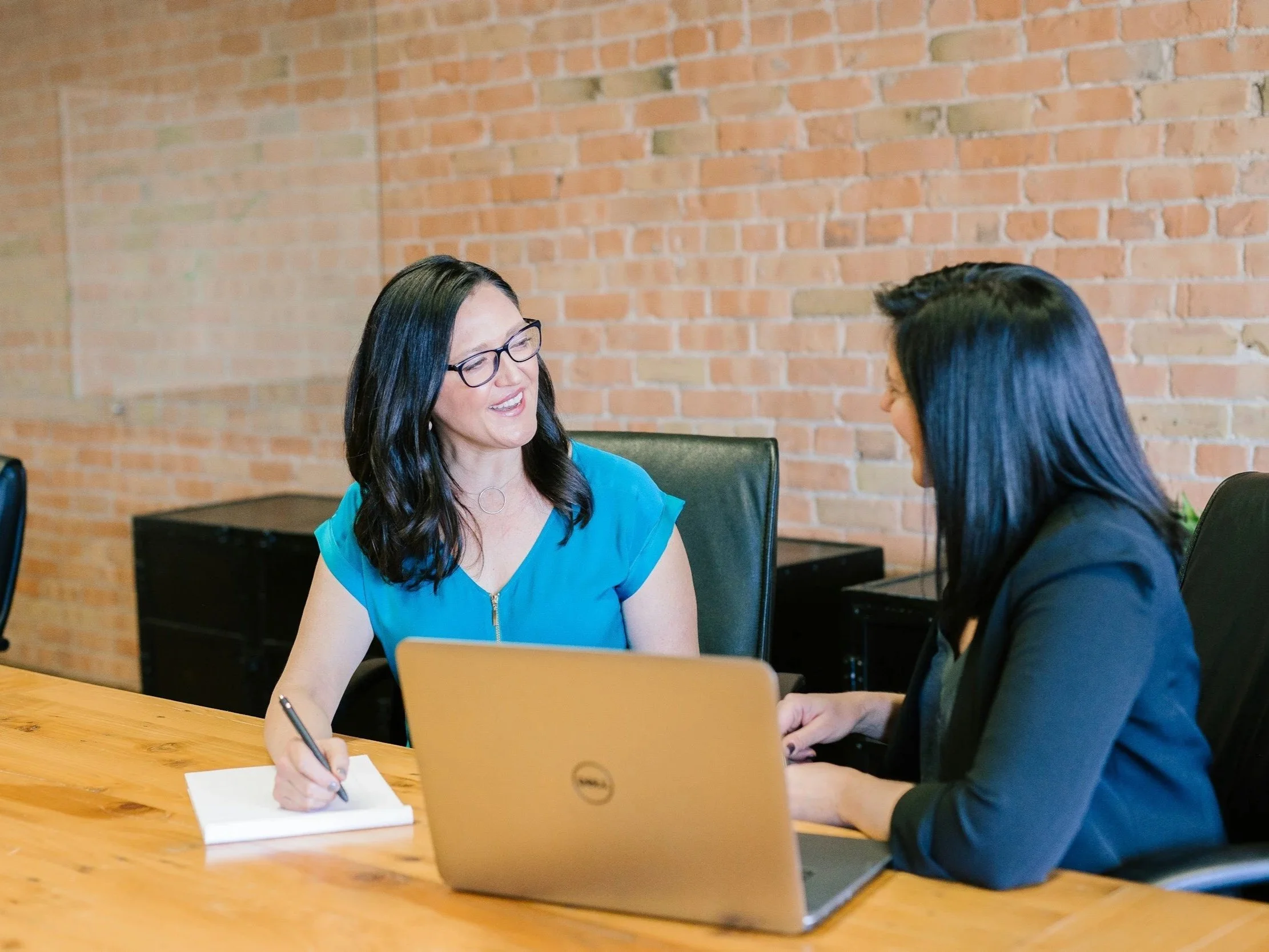 Two women sit at a desk, one wearing a dark suit coat and the other a teal blouse.