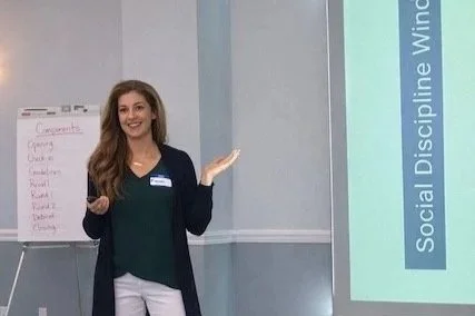 A woman with long brown hair presents a screen and stands next to an easel with circle components on it