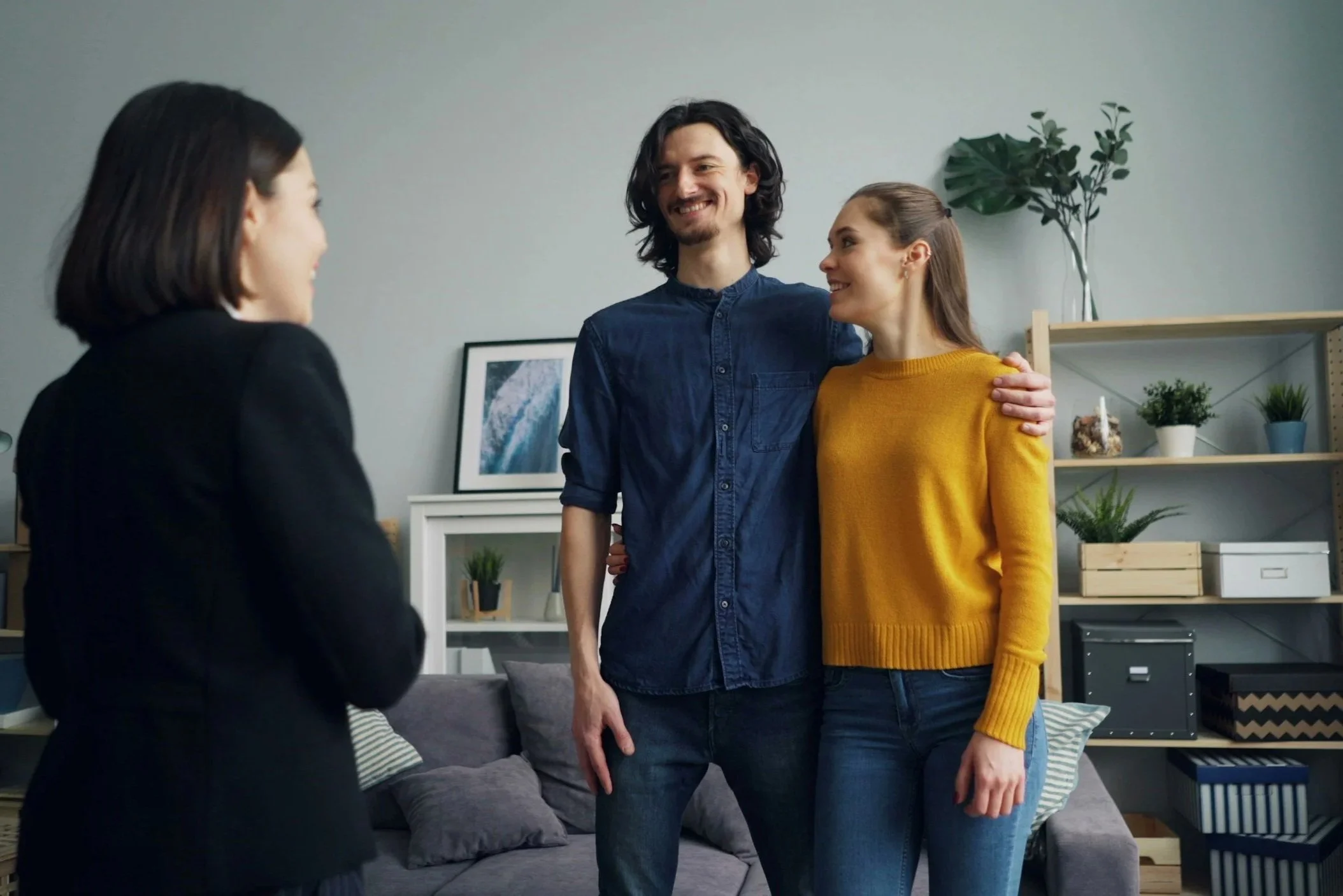 A man and woman embracing in a living room while a woman in a black blazer talks to them.