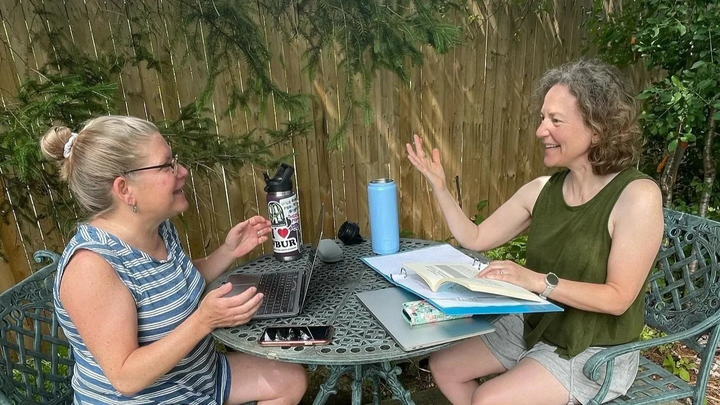 Beth and Rebecca sit outdoors at a small table with a laptop, notebooks, phones and water bottles. They are engaged in an animated discussion about PBIS.
