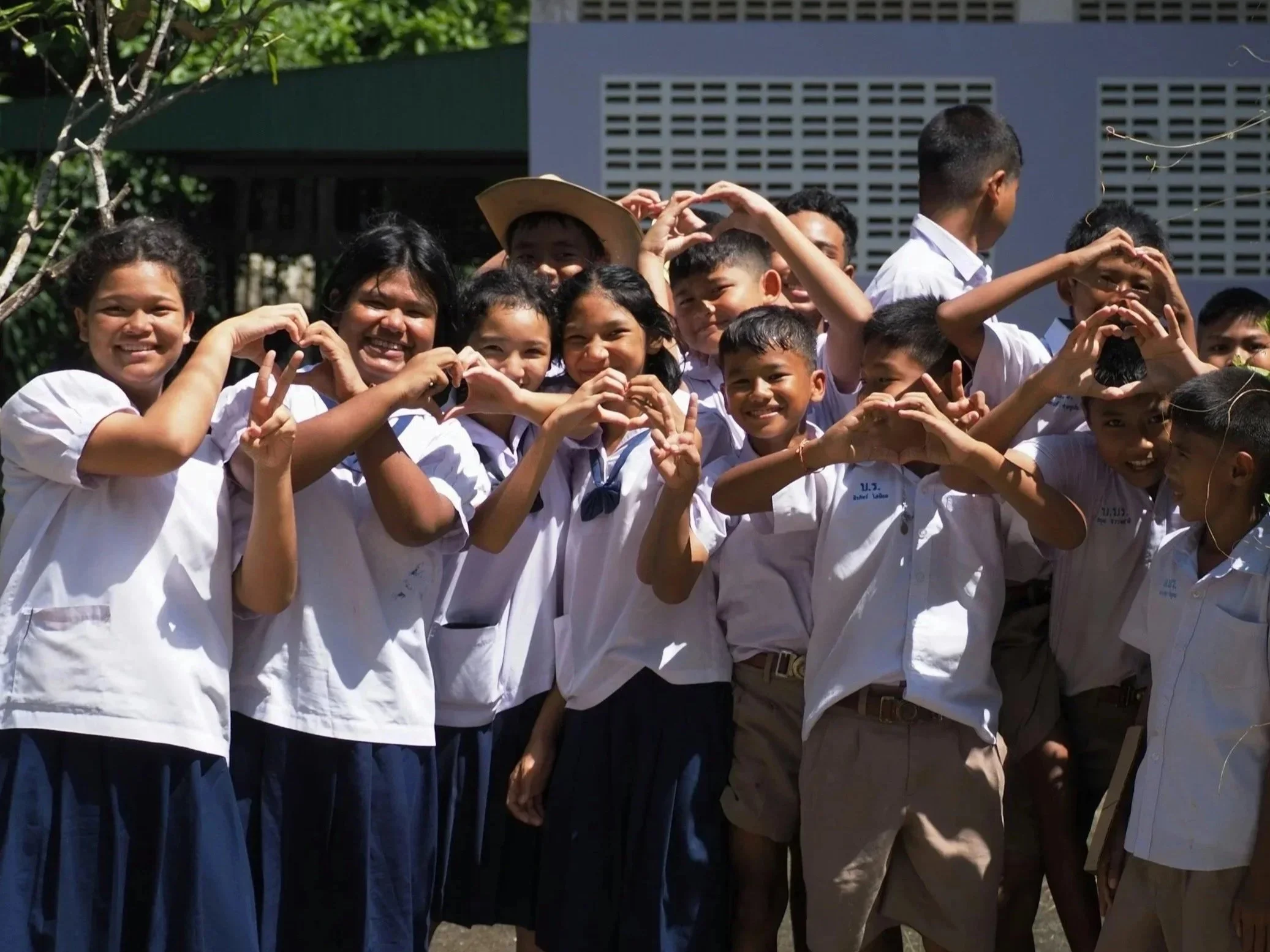 school aged children in uniform. The girls wear blue skirts and white shirts. The boys wear khaki pants and white shirts.  They are all smiling and holding their hands in a heart shape.