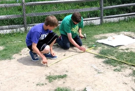 Two boys kneel outside on the ground, measuring a place for a garden bed. One holds the measuring tape and the other hammers a stake into the ground