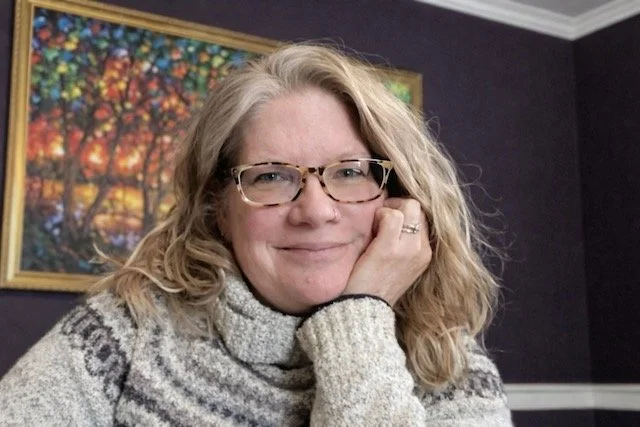 A woman with wavy blonde hair and glasses, smiling while resting her chin on her hand, wearing a gray knitted sweater, with a colorful landscape painting on the dark wall behind her.