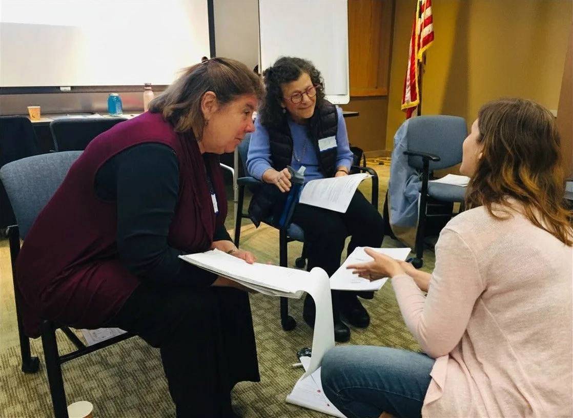 Two facilitators sit in chairs holding handouts while a participant sits on the floor facing them in a conversation.