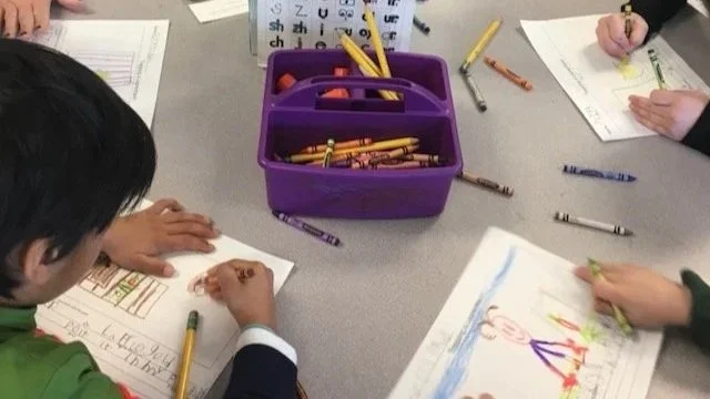 a closeup of students at a round table; the children are all drawing colorful pictures with crayons