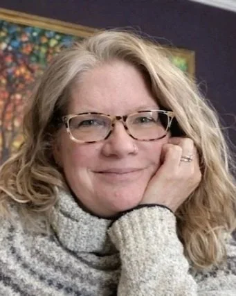 A blonde woman with glasses and a white sweater rests with her hand on her cheek
