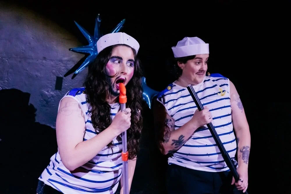 Two drag kings in sailor costumes perform on stage, one with blue star accessories and makeup, holding a microphone, and the other with tattoos, holding a microphone stand, against a dark background.