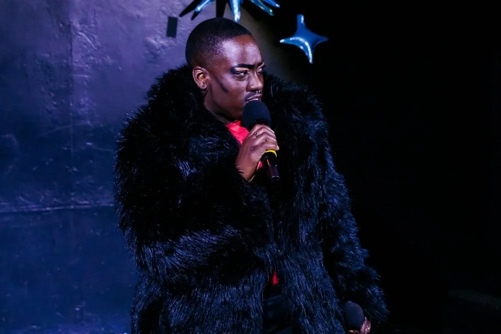 A person with short hair and dark skin holding a microphone on stage, wearing a black fur coat, with balloons and a dark background visible.