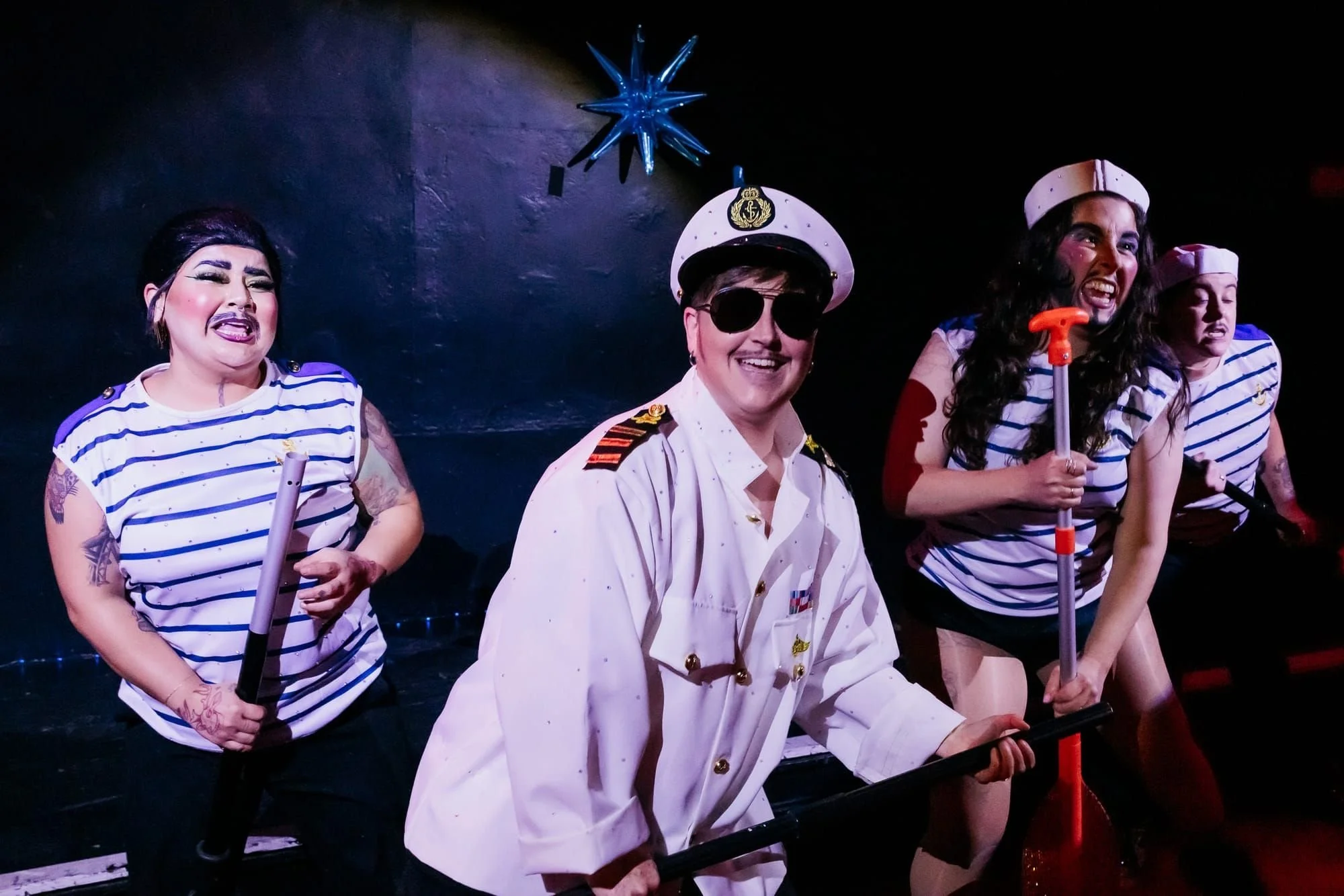 Four performers, three dressed as sailors and one as the captain in drag makeup and tattoos, standing on a dark stage with a blue star in the background.