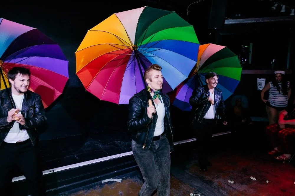 Three drag kings in leather jackets holding large rainbow-colored umbrellas, smiling and enjoying a night event indoors.