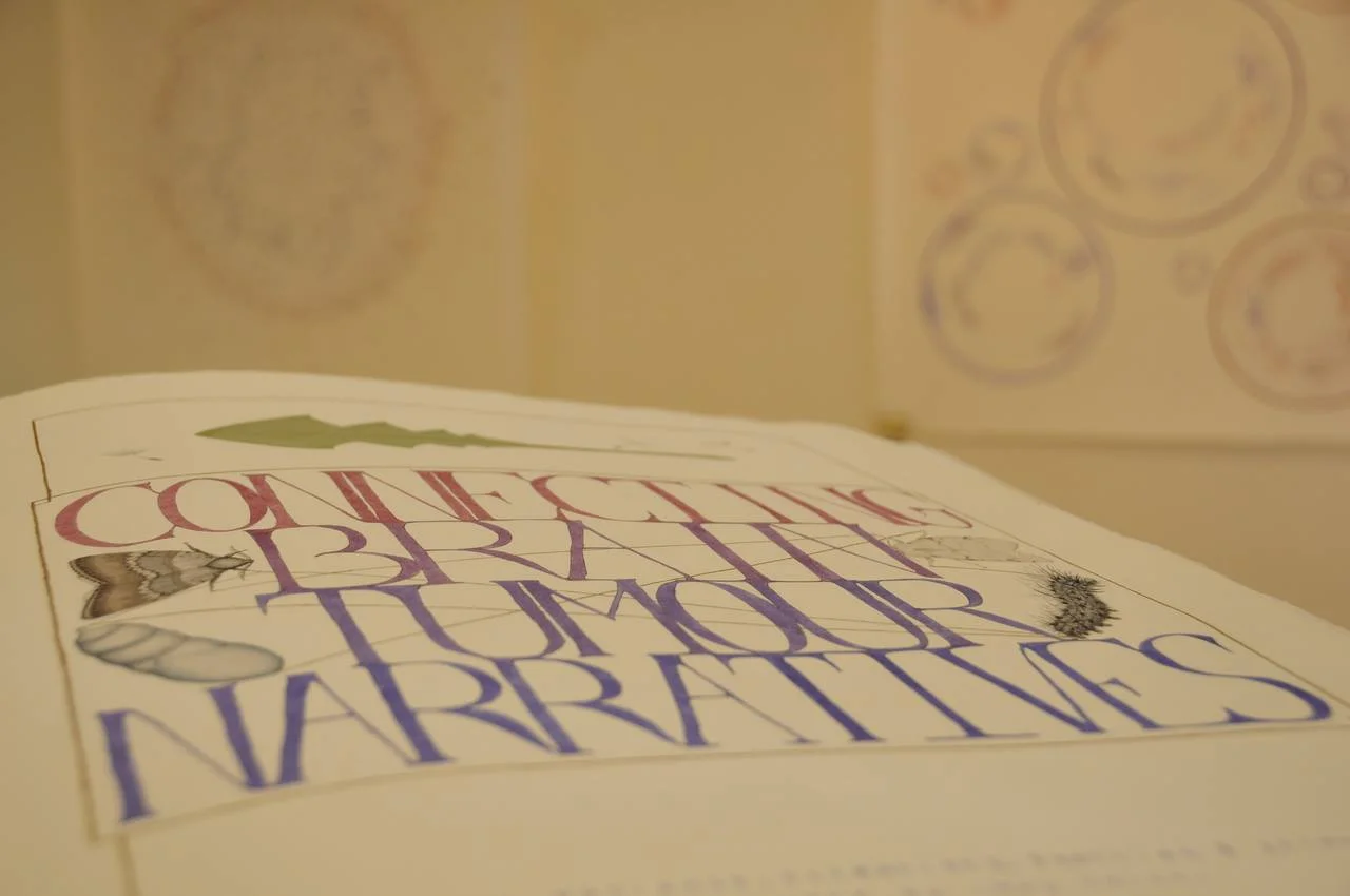 A giant handmade book at an art exhibition. The book is lying open showing the title page, which reads 'Connecting Brain Tumour Narratives" in shades through pink to purple.