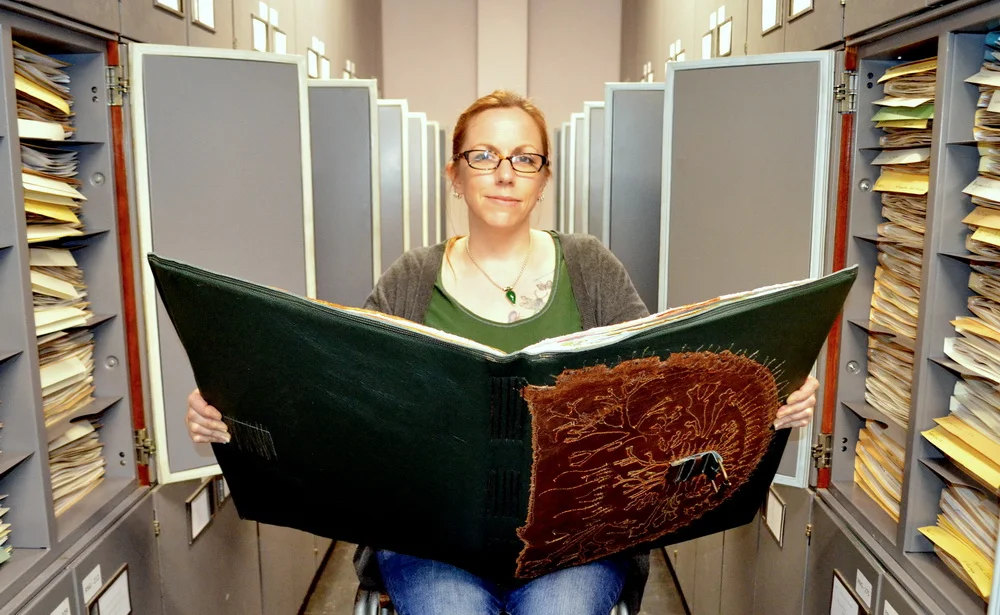 A person in a green tshirt, grey cardigan, and blue jeans, holding open a very large handmade book, in a herbarium (a collection of plant specimens.) Grey herbarium cupboards can be seen open behind the person, filled with yellow folders of dry plant specimens.