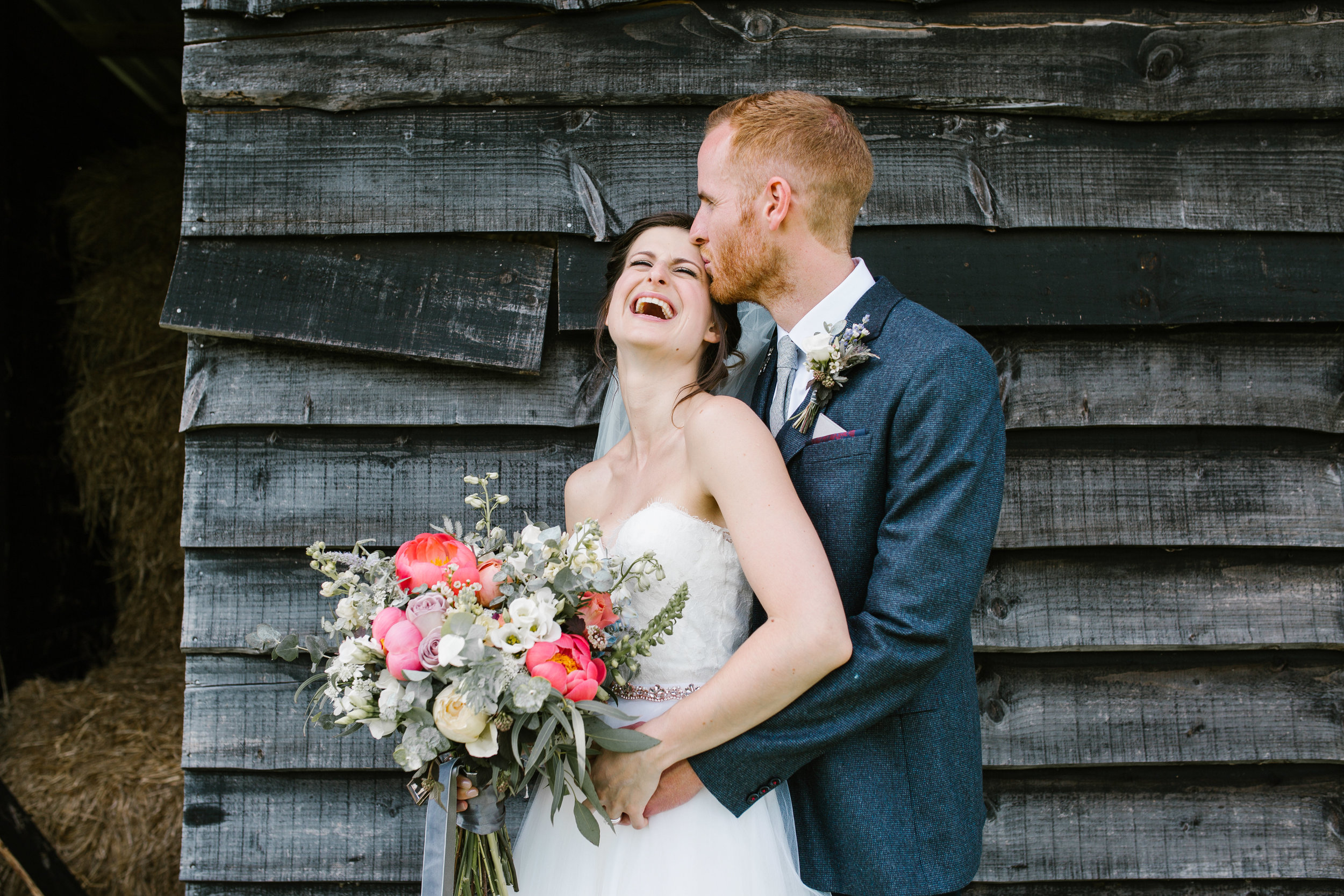 groom kisses his brides head as she laughs hysterically, standing in front of a rustic shed at upwaltham barns- west sussex