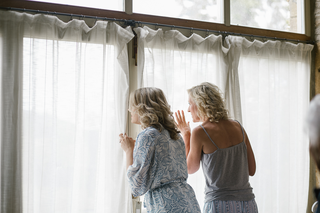 bride and mother of the bride peeking through the curtains as they watch the guests arrive at the Cotswolds wedding at Wildwood Blubell