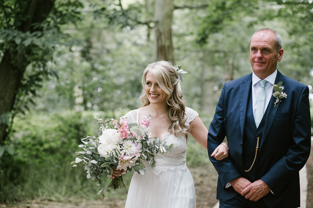 boho bride with father of the bride happily walking down the aisel in their outdoor woodland ceremony
