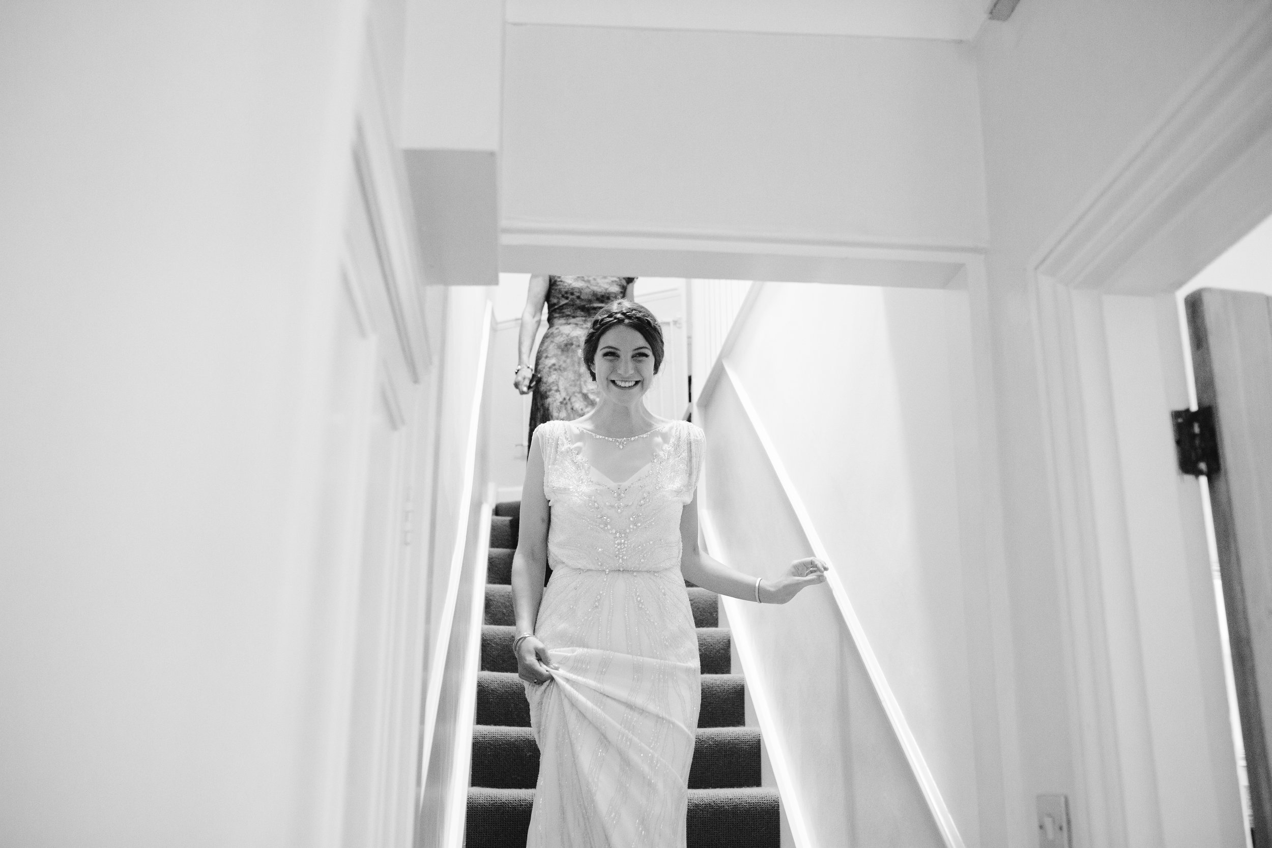 black and white photo of happy bride as she walks down the stairs towards her bridesmaids and dad