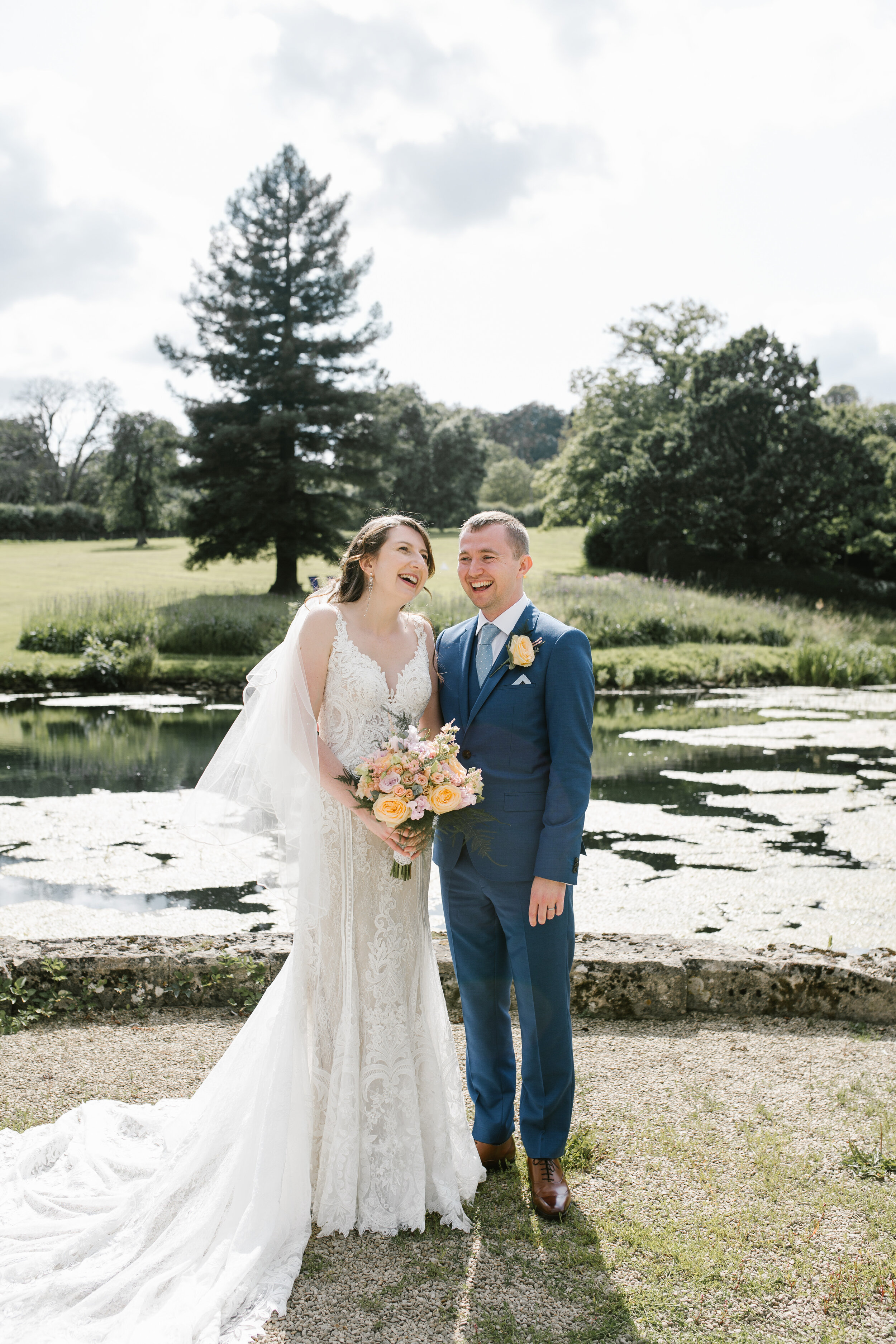 in the golden light the bride and groom laughing together in front of the lake at cowley manor
