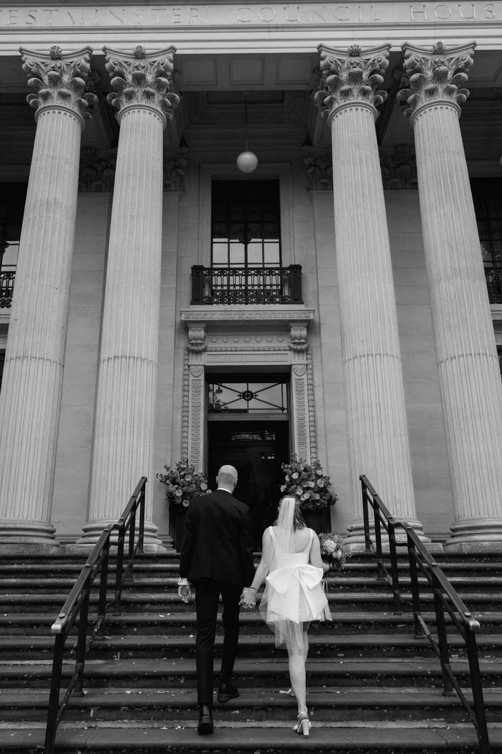 Black and white photo of the back of a bride and groom as they walk up the iconic stairs at Marylebone Town Hall, in London