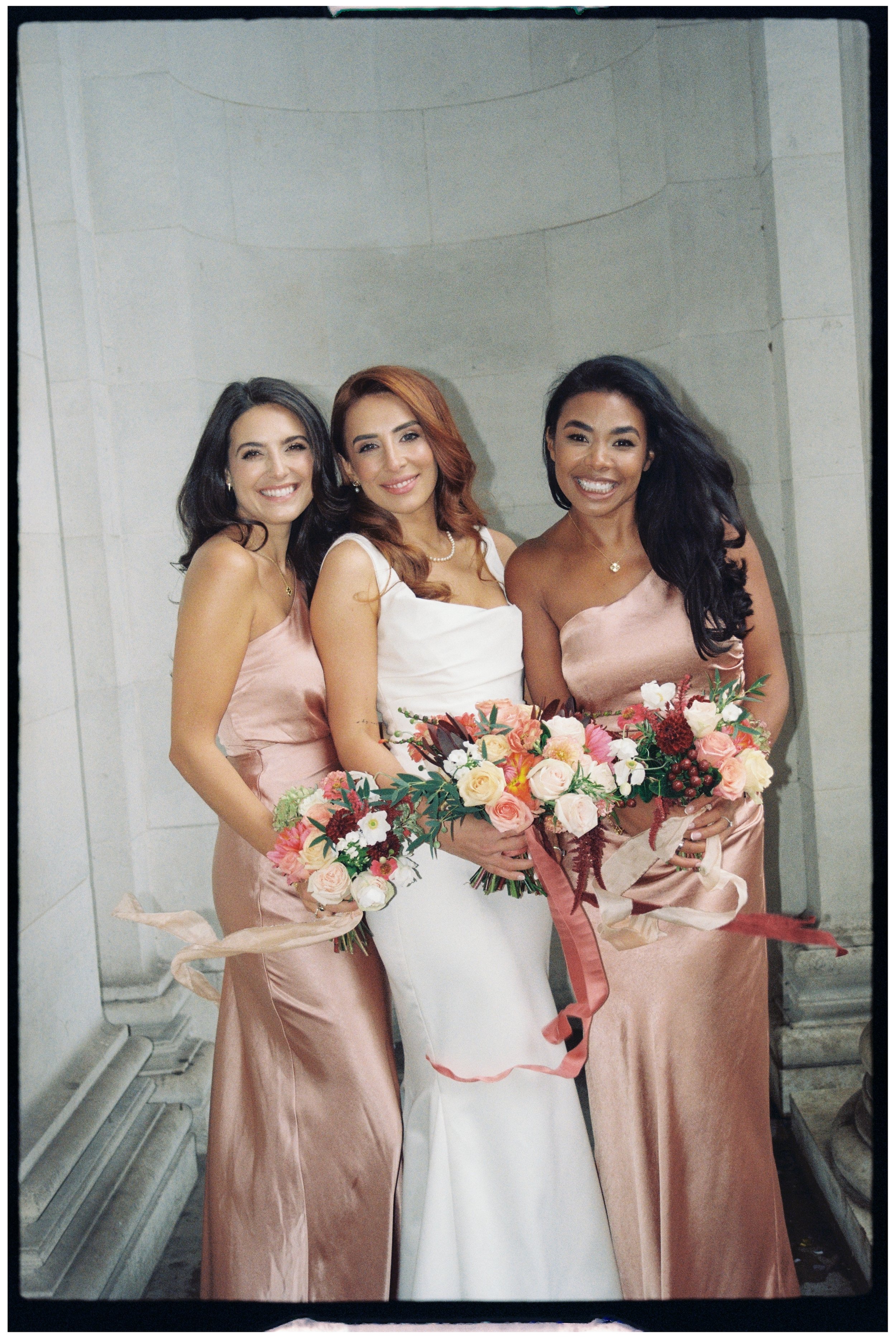 Bride with her Bridesmaids outside The Old Marylebone Town Hall smiling at the camera. Bridesmaids in peach silk dresses.