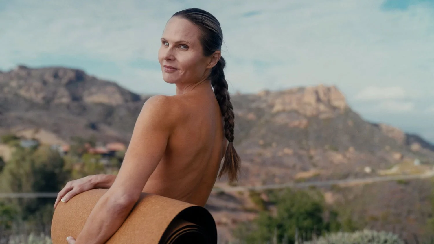 naked woman holding a rolled yoga mat looking at the camera