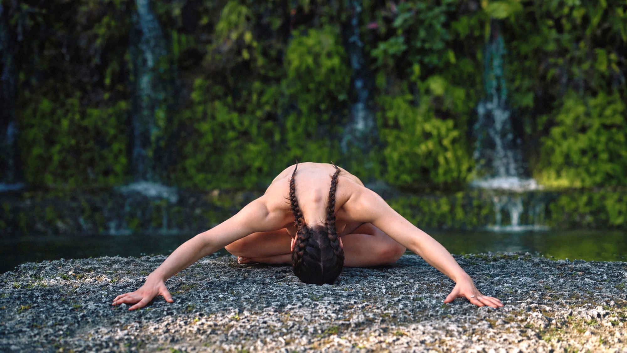 naked woman in cross-legged forward fold on rock with waterfalls in the back