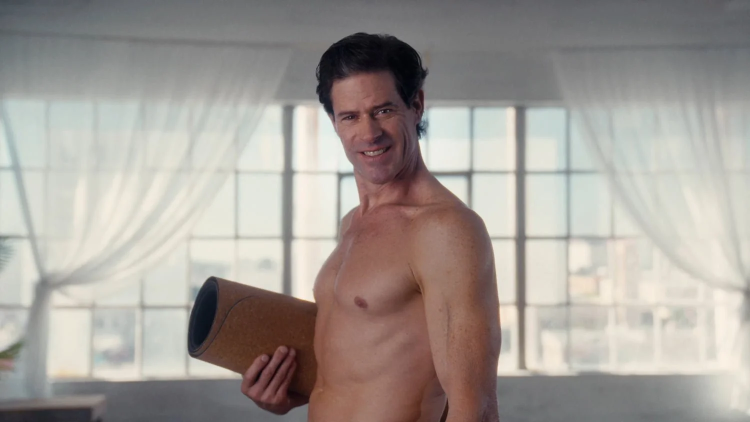 nude man smiling at camera holding a cork yoga mat