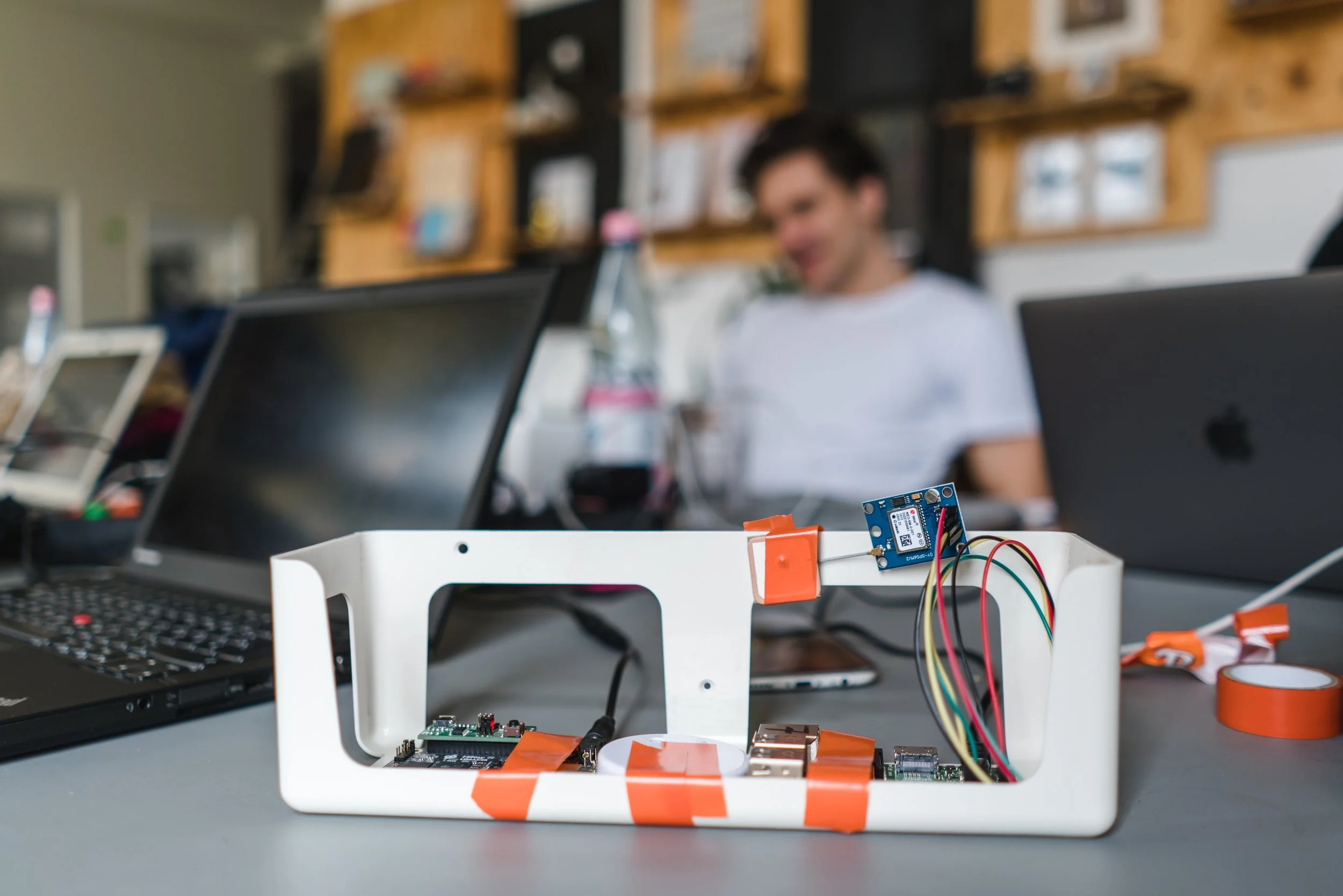 A prototype made with reused plastic components, and off-the-shelf electronic components, held together with electric tape. In the background, a team of engineers in front of their laptops at a table.