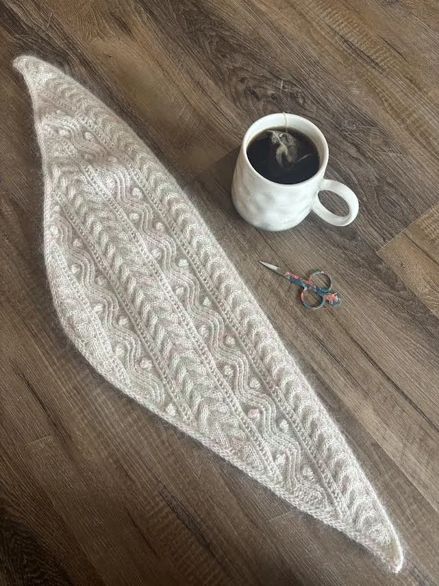 Photo of completed cabled shawl on wooden background with tea and scissors