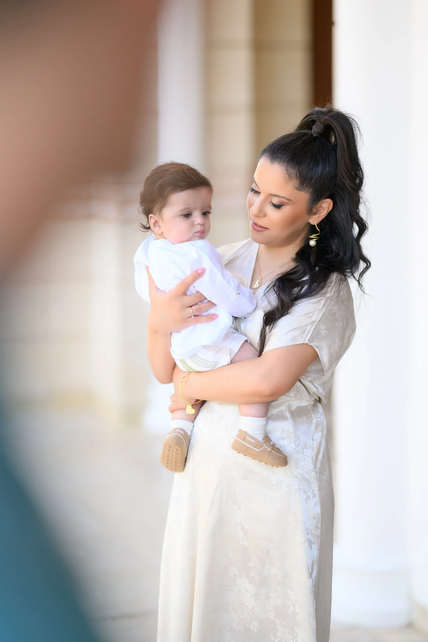 A woman with long dark hair in a high ponytail holding a young child in her arms, both dressed in light-colored clothing, standing outdoors in front of a building with columns.