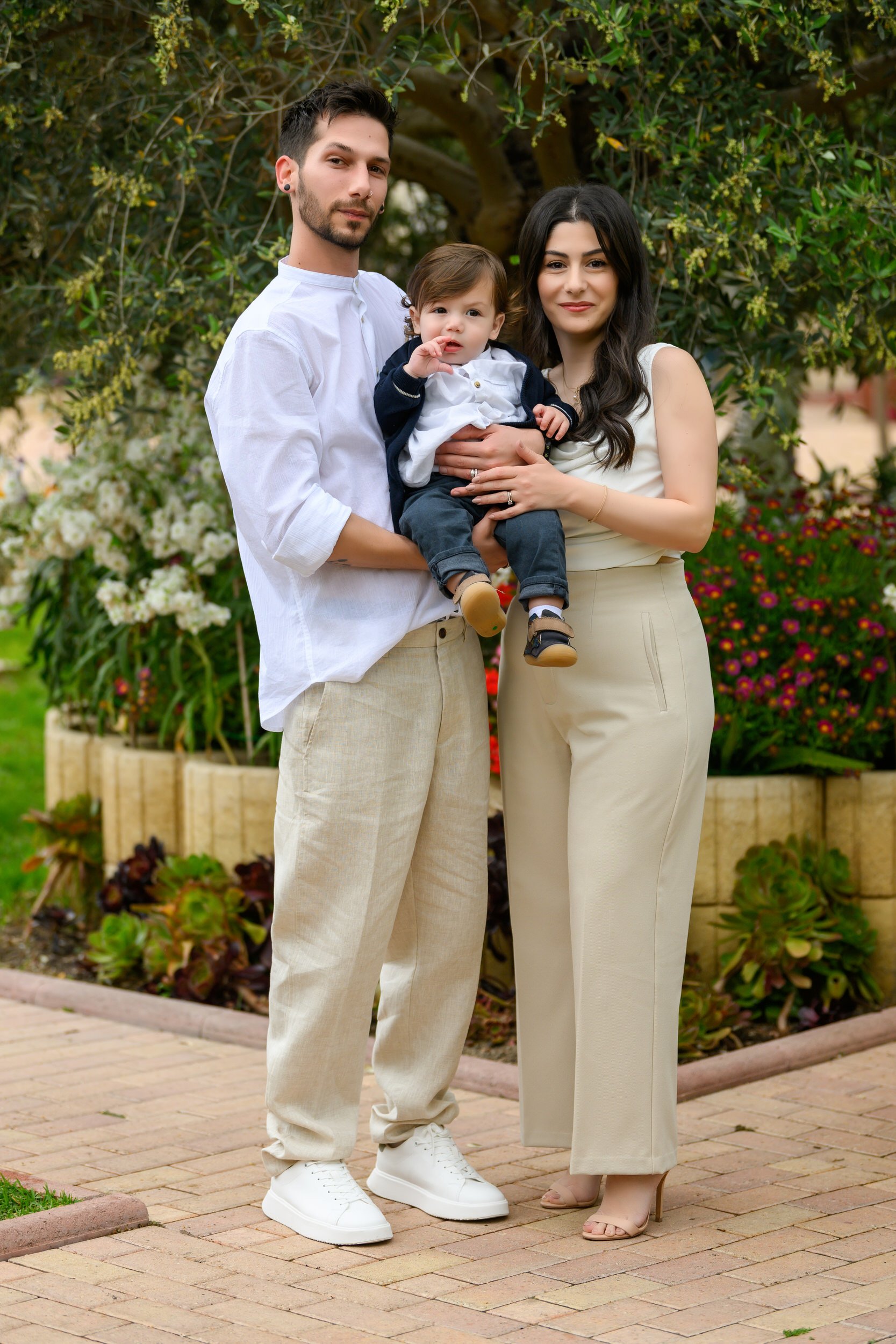 A family of three standing outdoors in front of a large tree and colorful flowers, with a man and woman holding a young child between them, all smiling