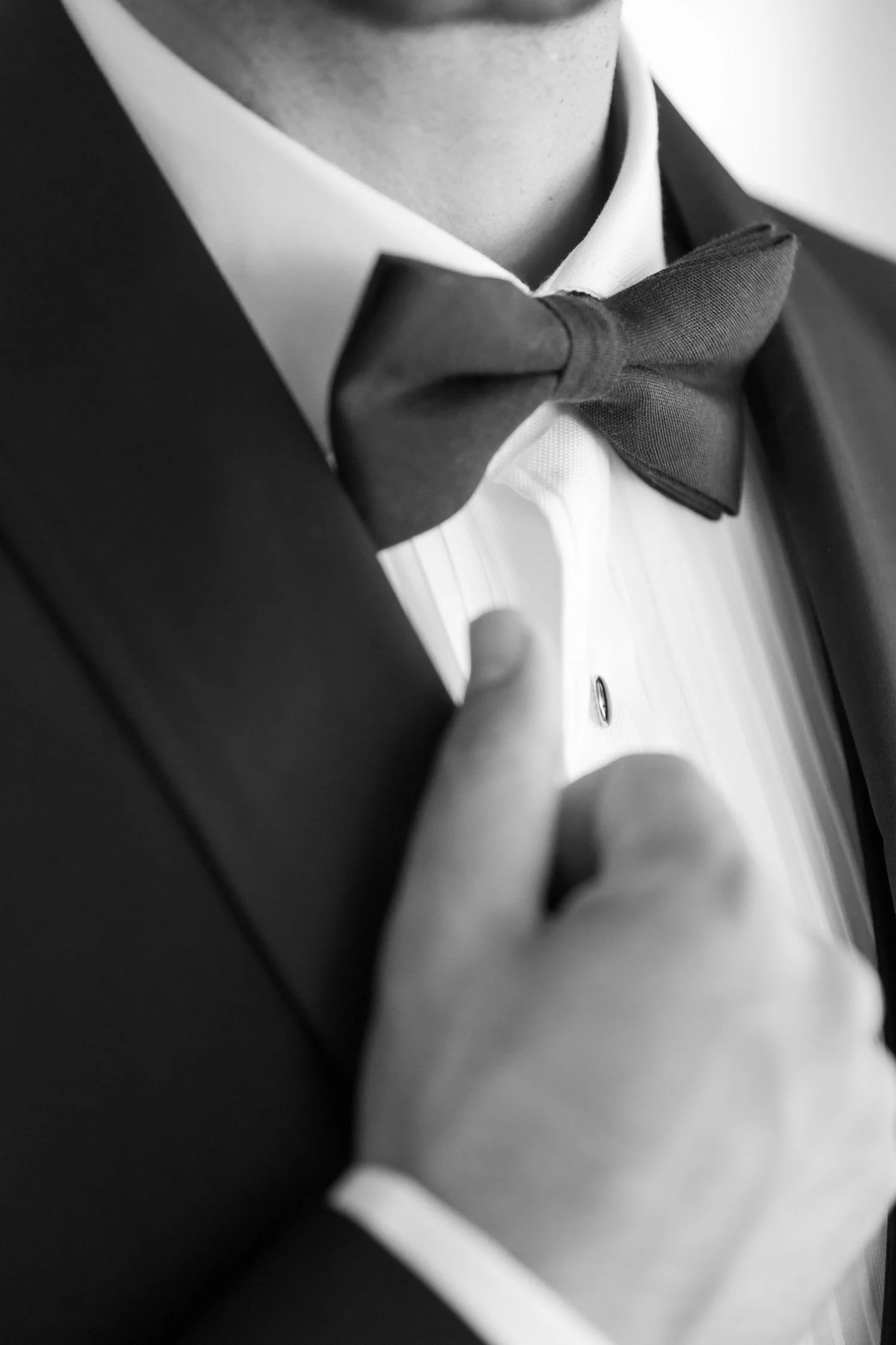 Close-up of a man adjusting his black bow tie while dressed in a tuxedo and white dress shirt.