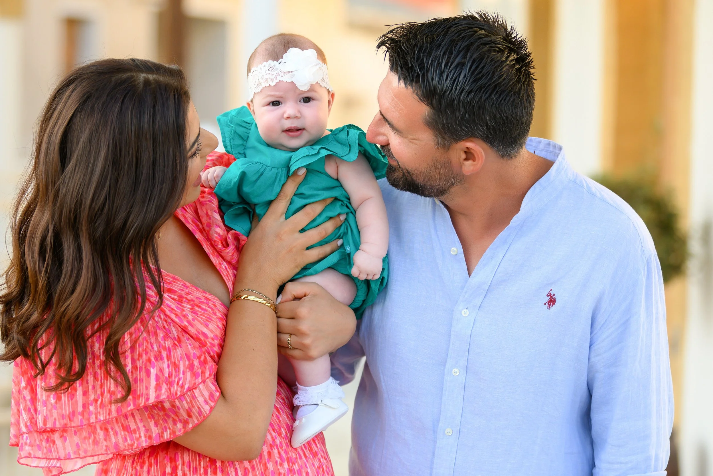 A family of three, a woman, a man, and a baby girl, sharing a moment indoors. The woman holds the baby, who wears a white headband and a teal dress, while the man smiles at her.