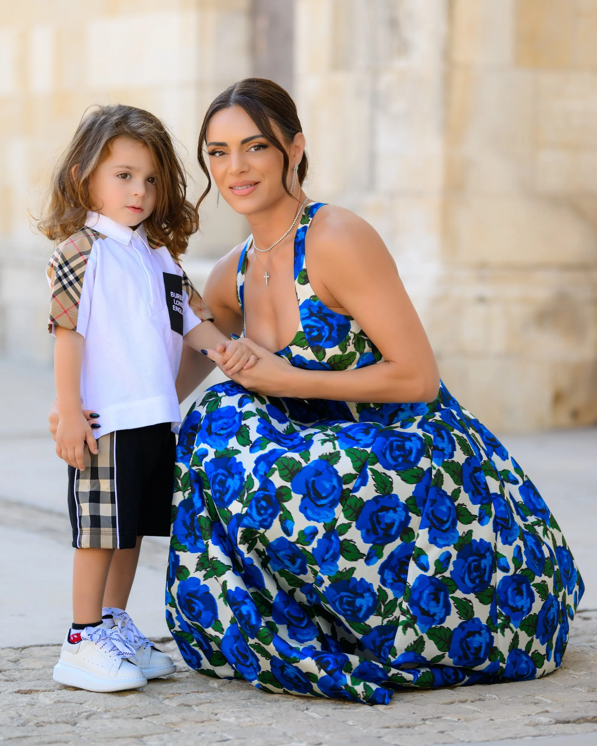 Woman in a blue floral dress kneeling next to a young girl in a white shirt and plaid shorts outdoors.
