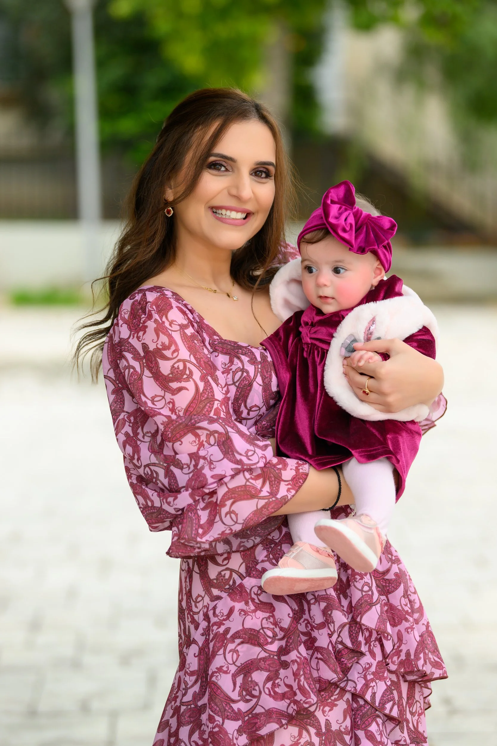 A smiling woman in pink and purple dress holding a young girl in a matching outfit with a large purple bow on her head, outdoors with green trees in the background.