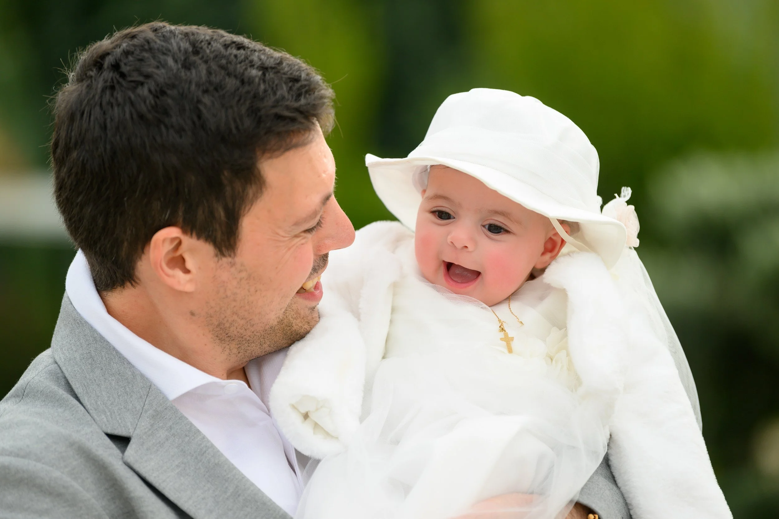 Man holding a baby girl dressed in white with a hat and cross necklace, smiling at each other outdoors.