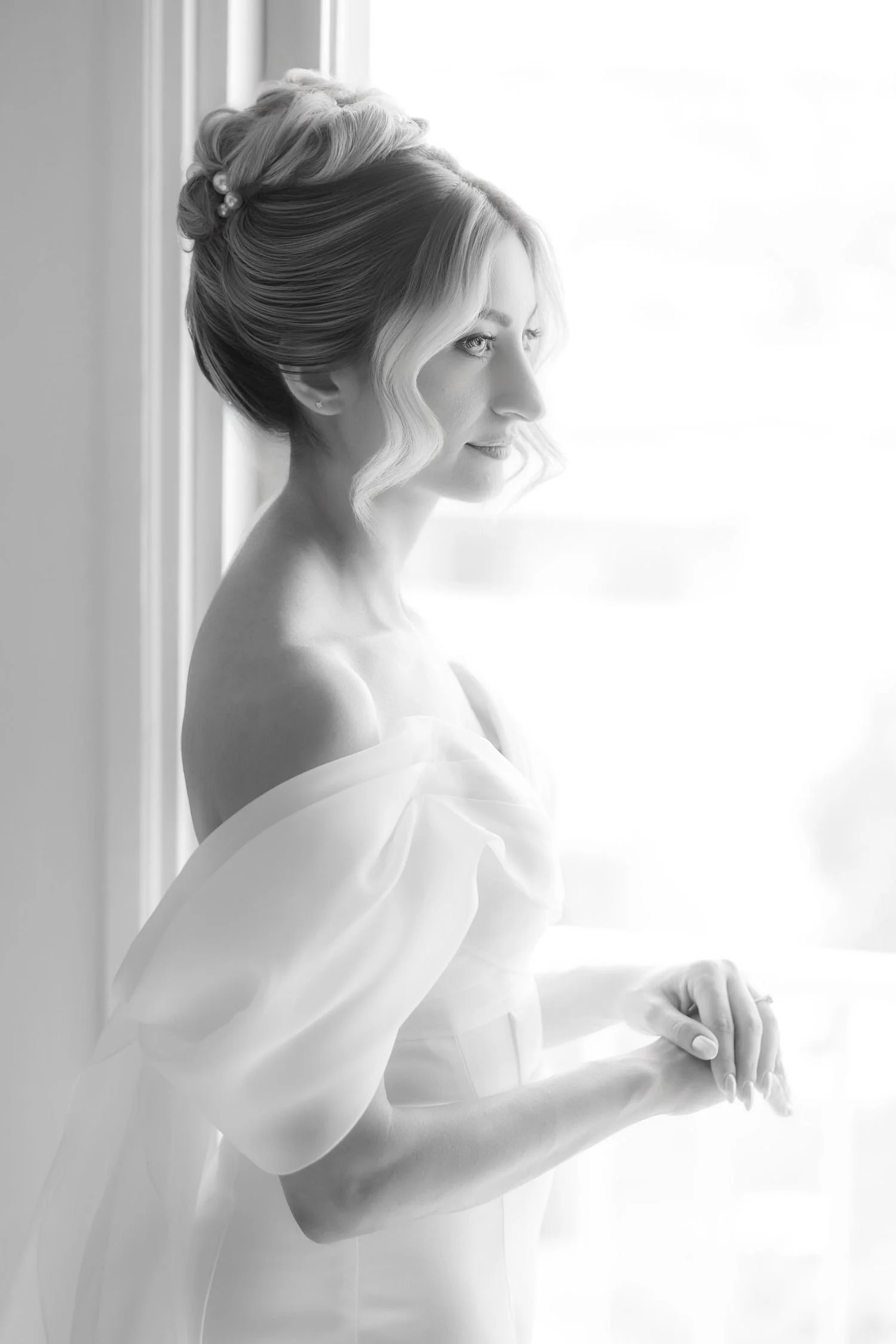 A black and white photograph of a woman with styled hair, looking out a window with soft lighting.