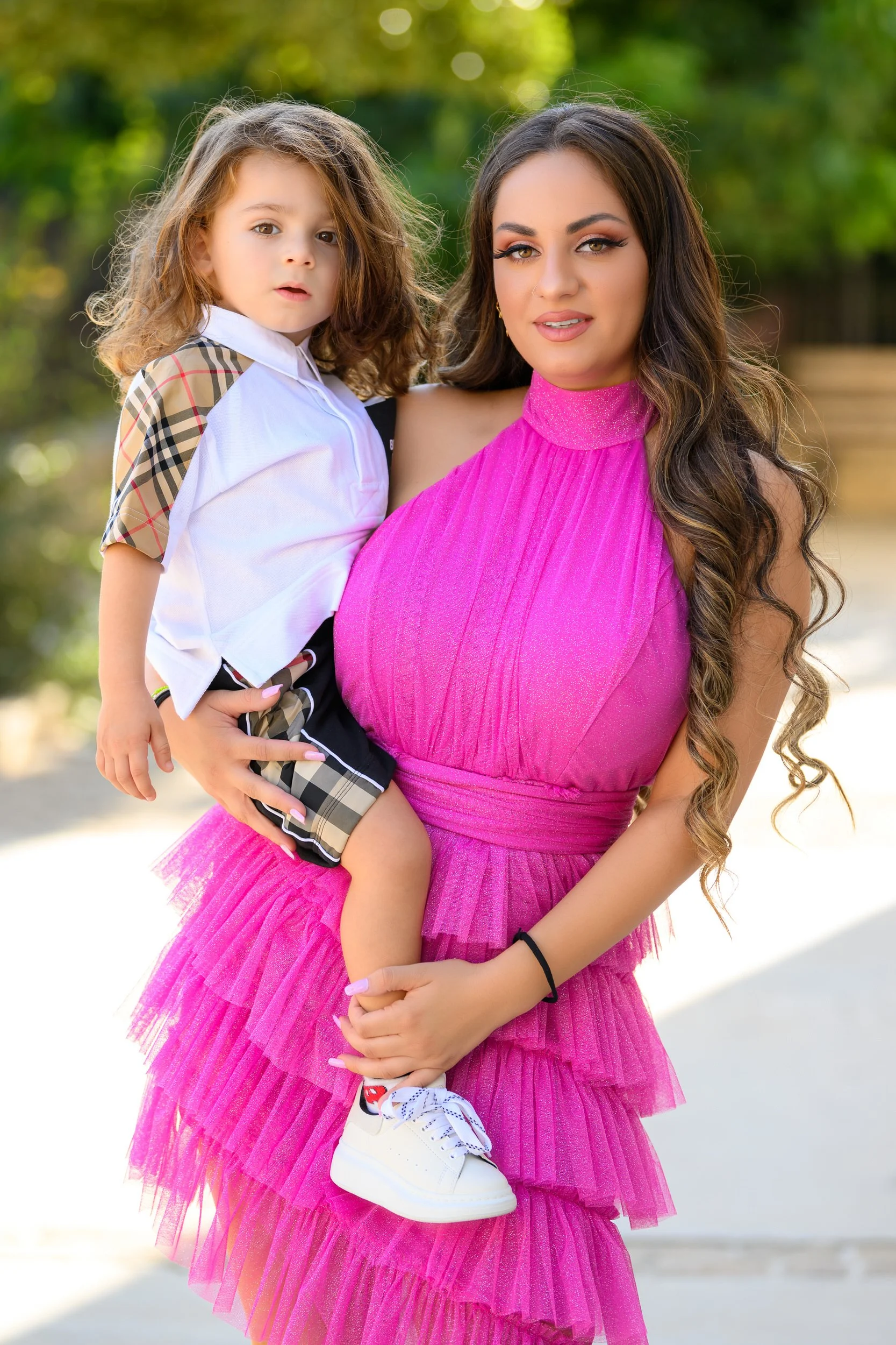 A woman in a vibrant pink dress holding a young girl in her arms outdoors with greenery in the background.